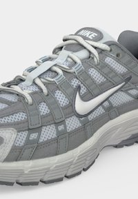 Γκρι και λευκό αθλητικό παπούτσι Nike με ύφασμα διχτυού, επενδεδυμένη γλώσσα και το λογότυπο Nike Swoosh στο πλάι και στη γλώσσα.