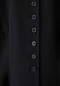 Gros plan sur un tissu noir avec une rangée verticale de six boutons ronds foncés cousus avec du fil noir.