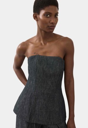 Kvinde med kort hår iført en stropløs, mørk denim corset-top, stående mod en ensfarvet lys baggrund.