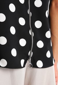 Svart blus med stora vita prickar, dragkedja fram, och ett lätt tyg med en slät yta.