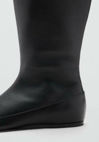 Bota de cuero negro hasta la rodilla con una textura suave, forma afinada y un tacón de cuña bajo y elegante. Presenta un diseño minimalista sin adornos.