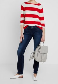 Röd- och vitrandig långärmad topp, mörkblå snäva jeans, vita platta skor och en ljusgrå ryggsäck. Enkelt, avslappnat design.