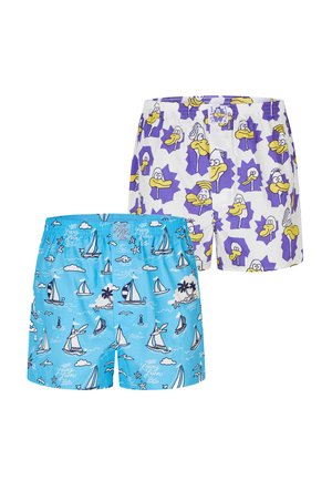 Muskelbepackter Mann mit geflochtenem Haar und Kinnbart steht frontseitig und trägt hellblaue Boxershorts mit weißen Segelboot- und Wolkenmotiven.