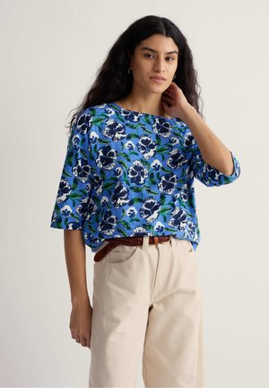 Femme aux longs cheveux noirs portant une chemise à fleurs bleues et un pantalon beige, une main touchant le cou, debout devant un fond uni.