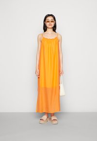 Vestido maxi laranja sem mangas com decote franzido, tecido leve, forma fluida e uma seção inferior transparente. Combinado com sandálias de plataforma bege.