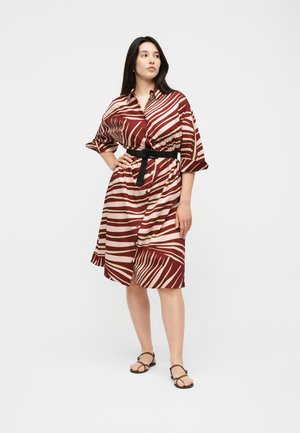 Donna in piedi con la mano sul fianco, indossa un vestito camicia con cintura a righe zebrate color bordeaux e crema e sandali neri su uno sfondo semplice.