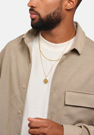 Goldfarbene, übereinander getragene Halsketten mit einem runden Anhänger und filigranen Details, getragen über einem weißen Hemd und einer hellbraunen, strukturierten Jacke.