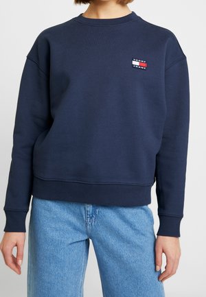 Femme portant un sweat-shirt Tommy Jeans bleu marine et un jean en denim bleu clair sur un fond clair uni.