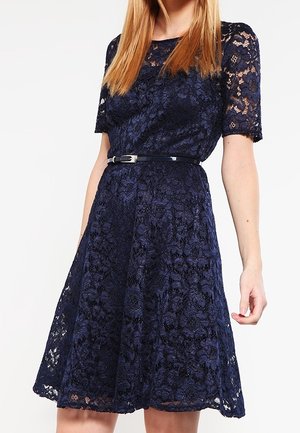 Kobieta ubrana w granatową koronkową sukienkę do kolan z krótkimi rękawami i cienkim czarnym paskiem w talii na białym tle.