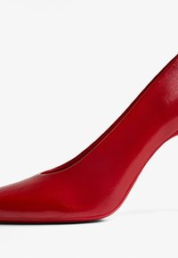 Cumpăna roșie cu toc înalt, fabricată din material sintetic lucios. Dispune de un vârf ascuțit și un toc subțire, curbat. Textură netedă cu detalii minime.