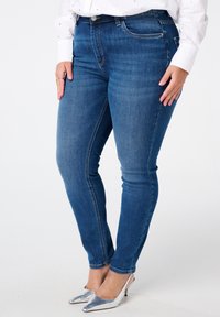 Jean en denim bleu skinny avec une texture lisse, présentant un design traditionnel à cinq poches, des accessoires en argent et une taille mi-haute.