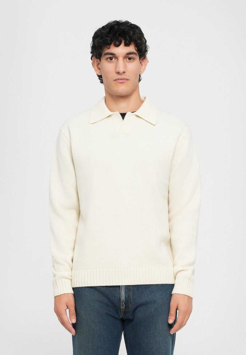 Suéter color crema con cuello tipo polo, puños y dobladillo acanalados. Hecho de un material de punto texturizado. Llevado con jeans azules.