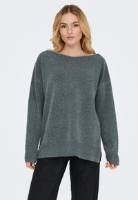 Prevelik temno zeleno moder pulover iz mehke pletene tkanine, z širokim izrezom v obliki čolna, rebrastimi manšetami in daljšo zadnjo obrobo.