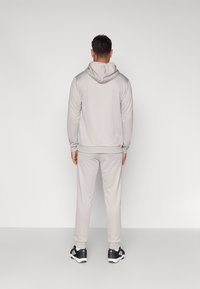 Ανοιχτό γκρι φούτερ με κουκούλα και αντίστοιχο παντελόνι jogger από μαλακό ύφασμα, με πλεκτά μανσέτες και τελείωμα, και κουκούλα με κορδόνι. Φοριούνται μαύρα αθλητικά παπούτσια.