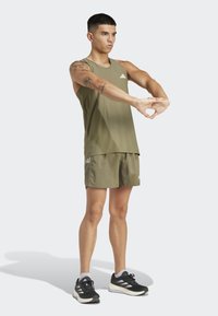 Green athletic tank top and shorts with three white stripes on shorts, worn with black running shoes. Model stretching arms outward.