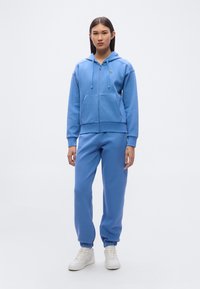 Ανοιχτό μπλε φούτερ με κουκούλα και κλείσιμο με φερμουάρ, συνδυασμένο με ταιριαστά παντελόνια φόρμας. Διαθέτει τσέπες μπροστά και ελαστικές μανσέτες. Φορεμένο με λευκά αθλητικά παπούτσια.