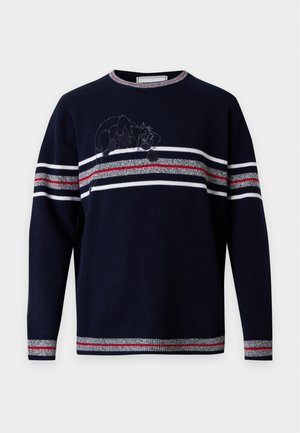 Homme portant un pull bleu marine à rayures rouges et blanches, pantalon beige, baskets bleues et blanches, debout devant un fond blanc uni.