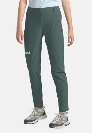 Persoon die donkergroene Jack Wolfskin wandelbroek en grijze wandelschoenen draagt, staand tegen een witte achtergrond.