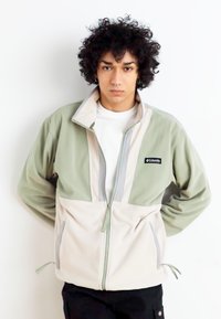 Giacca in pile leggera con zip, dal design bicolore; parte superiore in verde chiaro, parte inferiore in beige, con tasche laterali e cerniere a contrasto.