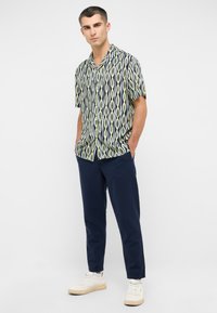 Koszula z krótkim rękawem w niebiesko-zielono-kremowy falisty wzór, zestawiona z granatowymi spodniami i białymi trampkami. Gładka tekstura materiału.