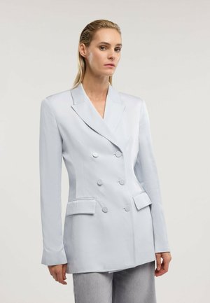 Frau mit zurückgekämmtem blondem Haar, die einen hellblauen zweireihigen Blazer und graue Hosen trägt, vor einem schlichten Hintergrund stehend.