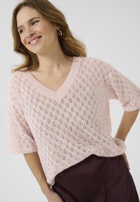 Pull léger rose en tricot avec un col en V, des manches courtes et un motif en dentelle. Texture douce avec une coupe décontractée, associé à un bas sombre.