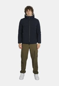 Giacca puffer blu navy con colletto alto e due tasche frontali con zip, abbinata a pantaloni cargo olive con multiple tasche e una zip.