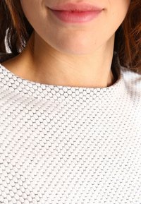 Top blanco con patrón hexagonal negro, con un cuello alto y una textura suave. La vista en primer plano se centra en el cuello y el área superior del pecho.