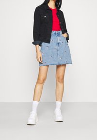 Falda de mezclilla con lavado azul claro, dobladillo deshilachado y patrones de corazones rojos bordados esparcidos, combinada con una chaqueta de mezclilla negra y zapatillas blancas.