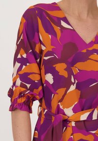 V-halsjurk met opvallende paarse, oranje en crèmekleurige bloemmotieven. Getextureerde pofmouwen met elastische manchetten en een bijpassende riem.