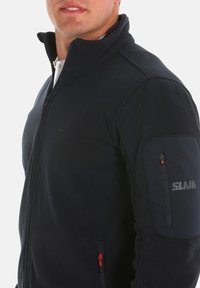 Navy-Fleecejacke mit hohem Kragen, Seiten- und Armtaschen mit Reißverschluss sowie Logodetail am Arm. Glatte Textur mit kontrastierenden Reißverschlüssen.