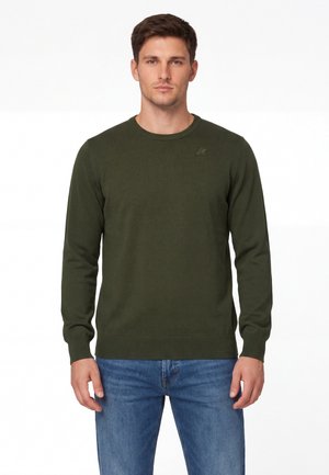 Młody mężczyzna ubrany w oliwkowo-zielony sweter z długim rękawem i niebieskie jeansy, stojący na tle jednolitego białego tła.