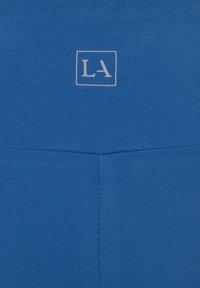 Blå leggings med ribbad textur. En liten logotyp med "LA" i vitt finns vid midjan. Enkelt design utan mönster.
