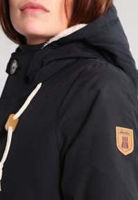 Marinblå huva jacka med en strukturerad yta, försedd med krämfärgade dragsnören och en tanfärgad broderad logotyp på ärmen.