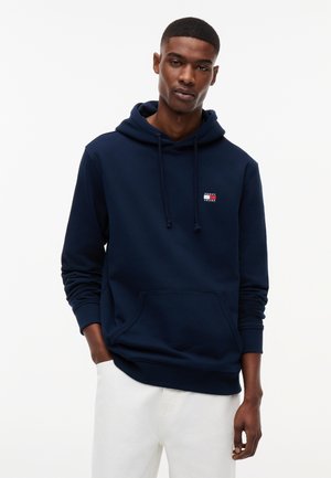 Νεαρός άνδρας που φοράει μπλε σκούρο φούτερ με κουκούλα, τσέπη μπροστά και λογότυπο Tommy Jeans, συνδυασμένο με λευκό παντελόνι, στέκεται σε απλό λευκό φόντο.