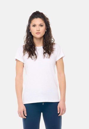 Giovane donna con lunghi capelli ricci che indossa una camicia bianca a maniche corte e pantaloni blu scuro, in piedi davanti a uno sfondo chiaro uniforme.