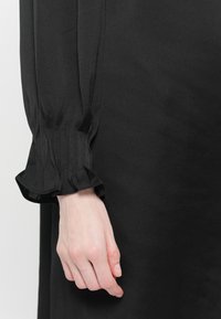 Svart kjole med lange, teksturerte ermer og volanger på mansjett. Stoffet ser glatt ut med en svak glans, som fremhever den strukturerte designen.