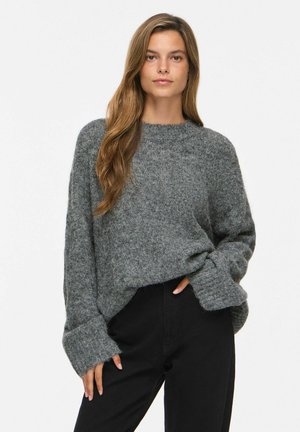 Femme aux longs cheveux bruns portant un pull en maille gris ample et un pantalon noir, debout avec une main dans la poche devant un fond uni.