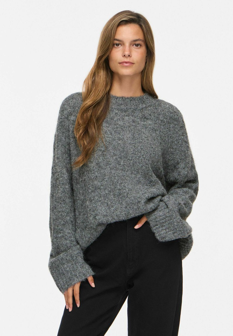 Femme aux longs cheveux bruns portant un pull en maille gris ample et un pantalon noir, debout avec une main dans la poche devant un fond uni.