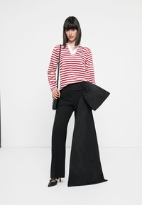 Frau mit glattem, dunklem Haar, die ein langärmliges rot-weiß gestreiftes Shirt, schwarze Hosen und High Heels trägt. Sie hält einen großen schwarzen Stoff und eine kleine schwarze Handtasche.