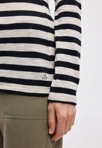 Camisa de manga longa às riscas em azul-marinho e creme, feita de um tecido macio. Apresenta um pequeno logo bordado perto da bainha. Combinada com calças verde-oliva.