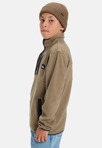 Veste en polaire beige avec un col haut, une patte de boutonnage noire, des poches latérales et un petit patch logo ; associée à un bonnet en tricot marron.
