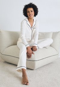 Krämfärgad pyjamasett med marinblå piping, bestående av en skjorta med knappar och långa byxor, tillverkad av mjukt tyg, sittande på en strukturerad loungefåtölj.