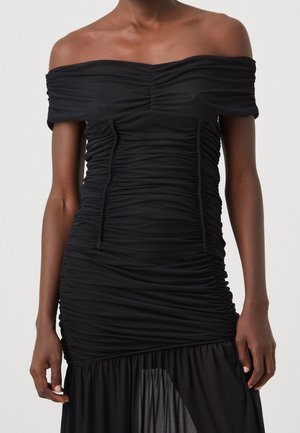 Personne portant une robe noire épaules dénudées avec des détails froncés et un tissu transparent en bas de la jupe, sur un fond uni.