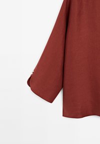 Détail de la manche d'un chemisier en lin couleur rouille avec trois petits boutons blancs près du poignet sur un fond blanc uni.