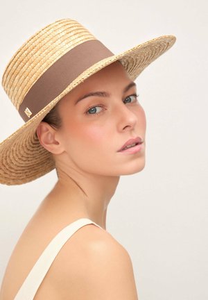 Mlada ženska s svetlo kožo in modrimi očmi, ki nosi širok rob klobuka iz slame z rjavim trakom, obrnjena čez ramo.