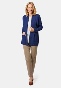 Cardigan bleu marine à devant ouvert, avec des poignets côtelés et deux poches avant, associé à un pantalon beige et des talons marron. Texture lisse.