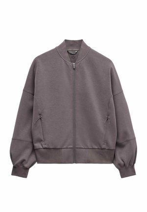 Kvinde iført oversized mauve lynlåstrøje med ribbede ærmekanter og matchende ribbede bukser, med sorte solbriller, poserer med hænderne på hofterne.