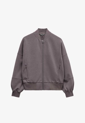 Kvinde iført oversized mauve lynlåstrøje med ribbede ærmekanter og matchende ribbede bukser, med sorte solbriller, poserer med hænderne på hofterne.