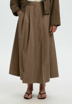 Kvinne iført en lang, taupe plissèert skjørt kombinert med svarte sandaler med remmer og en matchende taupe jakke over en hvit topp.